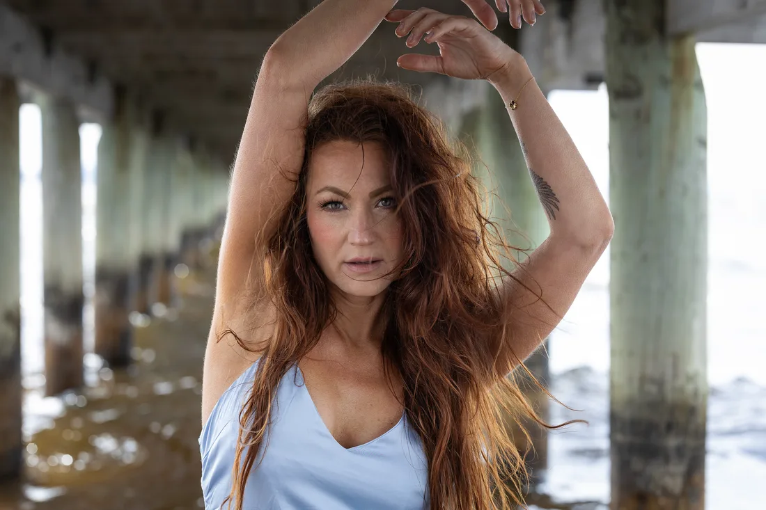 Woman in a pale blue dress under a wooden pier, arms raised, auburn hair filling the frame