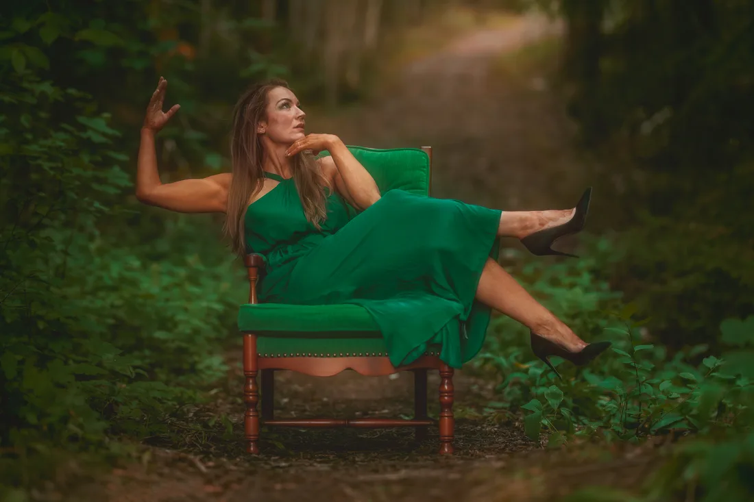 Woman in a long green dress seated in a green armchair on a forest path