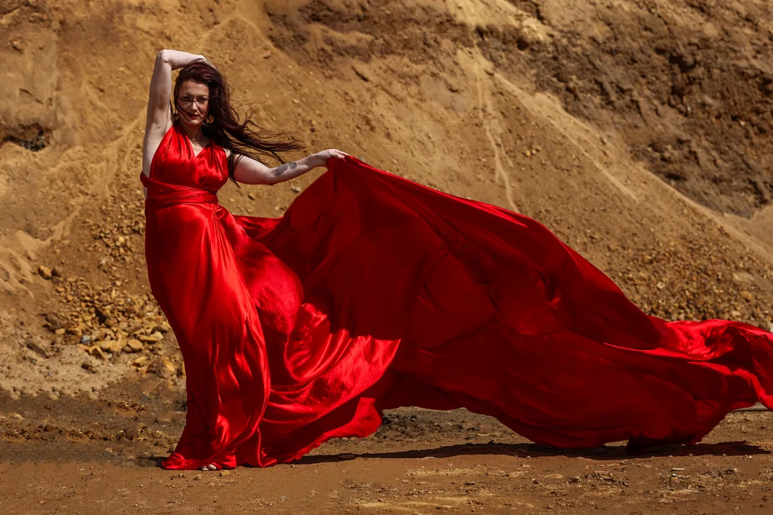 Woman in a red gown lifting the fabric into the wind against a wall of sand