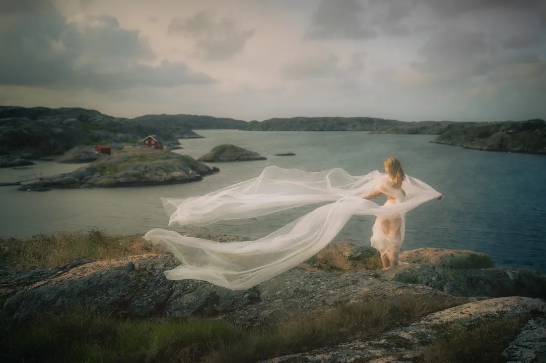 Figure on a rocky shore seen from behind, a long white veil blowing across a coastal inlet